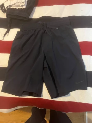 Svarta träningsshorts från Nike - Snygga svarta träningsshorts från Nike med snörning i midjan och två sidofickor. Perfekta för gymmet eller löprundan. Klassisk Nike-logga på benet och bekväm passform.