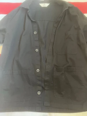 Svart overshirt med knappar H&m - Stilren svart overshirt med klassisk krage och tre stora fickor framtill. Skjortan har knäppning med fyra knappar och är tillverkad i ett mjukt bomullstyg. Perfekt att bära öppen eller stängd för en avslappnad look.