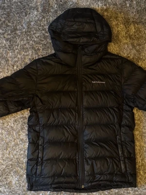 Svart dunjacka från Peak Performance - Snygg mörkblå dunjacka från Peak Performance med huva och dragkedja framtill. Jackan har en puffig design med horisontella sömmar och diskret logga på bröstet. Perfekt för kalla vinterdagar och riktigt varm tack vare dunfyllningen. Använd en vinter inget i min stil längre. (Storlek XL passar mer som L/M) Nåt märke vid dragkedjan annars är den nästan fläckfri. (Nypris för två år sedan 2300kr)