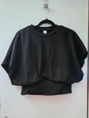 Svart croppad topp med vida ärmar - Säljer en svart croppad topp med bred resår i midjan och vida, korta ärmar. Toppen har rund halsringning och är gjord i ett mjukt, ribbat material som ger en avslappnad men trendig look.