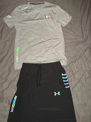 Under Armour träningsset grå/svart - Snyggt träningsset från Under Armour med en grå t-shirt och svarta shorts. Båda plaggen har färgglada loggor och detaljer i regnbågsfärger. T-shirten har rund hals och kort ärm, shortsen har dragsko i midjan och sidofickor. Perfekt för gym eller löpning.