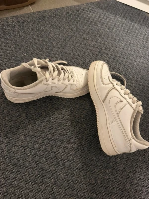 Vita låga sneakers i skinn - Säljer ett par klassiska vita sneakers i lågt modell med snörning och perforerade detaljer på tån. Skorna har en tjock sula och är tillverkade i skinn med vita sömmar och diskret logga på sidan. Perfekta för en clean och stilren look.