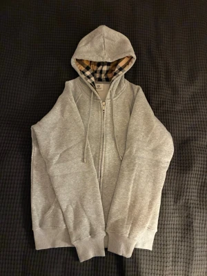 Burberry hoodie - Säljer en stilren grå hoodie med dragkedja och huva. Insidan av huvan har ett klassiskt beige, svart och vitt rutigt mönster. Hoodien har ribbade muddar vid ärmslut och nederkant samt dragsko i huvan. Perfekt för en avslappnad och trendig look.