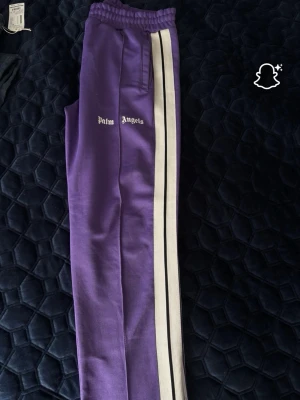 Lila Palm Angels mjukisbyxor - Säljer ett par lila mjukisbyxor från Palm Angels med vita och svarta ränder längs sidorna. Byxorna har resår i midjan, raka ben och fickor på sidorna. Perfekta för en chill och trendig look.