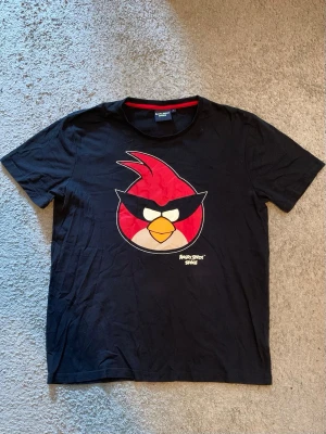 Svart Angry Birds Space t-shirt - Cool svart t-shirt med stort Angry Birds Space-tryck på bröstet. T-shirten har rund hals med röd detalj på insidan och är tillverkad i mjuk bomull. Perfekt för dig som gillar gaming och vill sticka ut med en ikonisk design.