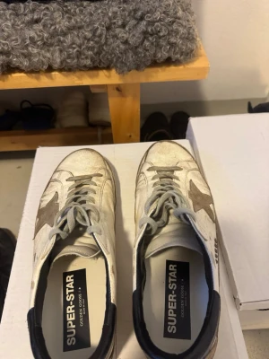 Vita Golden Goose Super-Star sneakers - Säljer ett par vita Golden Goose Super-Star sneakers med grå stjärndetalj på sidan och svarta detaljer vid hälen. Skorna har klassisk låg profil, snörning och sula i gummi. Ovandelen är i läder och mocka för en cool, avslappnad look. De är använda och de finns tyvärr större defekter på baksidan som man ser och även mindre på framsidan! Sjukt bekväma och snygga skor!