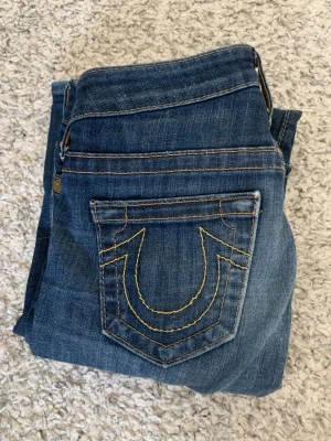 True Religon bootcut blå jeans med detaljerade sömmar - Säljer ett par klassiska blå jeans med tydliga kontrastsömmar och en unik bakficka med gul brodyr. Jeansen har normal midja, lite raka/flare ben och detaljer som metallknappar och flagga på insidan. Perfekta till en avslappnad streetstil.