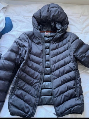Svart dunjacka med huva - Snygg svart dunjacka med huva och quiltade sömmar. Jackan har dragkedja framtill, två sidofickor och orange detaljer på insidan. Perfekt för kalla vinterdagar och ger en sportig look.