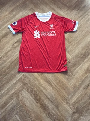 Liverpool matchtröja röd Nike - Säljer en röd Liverpool matchtröja från Nike med vita detaljer och Premier League-märke på ärmen. Tröjan har klubbmärke och sponsortryck på bröstet samt rund halsringning. Perfekt för fotbollsfans som vill visa sitt stöd på matchdagar. Storlek xxl men sitter som xl/L.  Oanvänd 