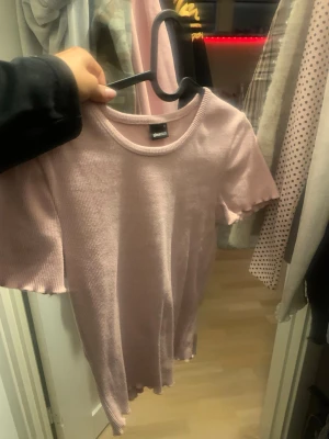 Ljusrosa  topp från Gina Tricot - Supersöt ljusrosa  topp från Gina Tricot. Modellen har korta ärmar, rund halsringning och vågiga kanter vid ärmslut och nederkant. Perfekt att styla med jeans eller kjol för en chill look.