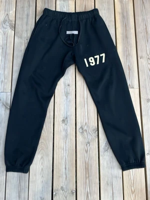 Svarta Essentials mjukisbyxor 1977 - Svarta mjukisbyxor från ESSENTIALS Fear of God med trycket '1977' på vänster lår. Byxorna har resår i midjan med dragsko och muddar vid bensluten. Perfekta för en avslappnad och trendig streetwear-look.