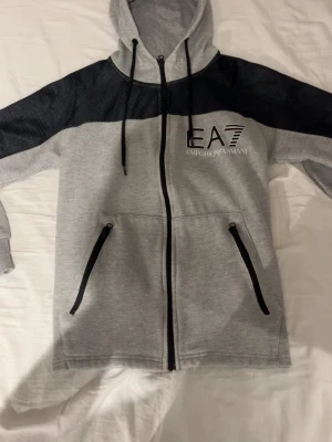 EA7 hoodie - Snygg grå hoodie från EA7 Emporio Armani med svarta meshpartier över axlarna, dragkedja framtill och två fickor med svarta dragkedjor. Stor logga på bröstet och justerbar huva med snören. Perfekt för en sportig och avslappnad stil.