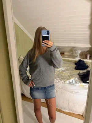 Grå stickad tröja med v-ringning - Mysig grå stickad tröja med bred v-ringning som kan bäras off-shoulder. Tröjan har långa ärmar och ribbade muddar vid ärmslut och nederkant. Perfekt till jeans eller kjol för en avslappnad look.