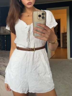 Vit broderad jumpsuit  - Supersöt vit jumpsuit med broderade detaljer och korta ärmar. Den har en feminin knytning vid bysten. Perfekt för sommardagar och enkel att styla med accessoarer.