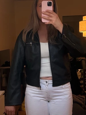 Svart skinnjacka med dragkedjor - Snygg svart skinnjacka med klassisk krage och dragkedjor vid ärmslut och bröstfick. Jackan har en rak passform och elastiska detaljer i midjan för en bekväm look. Perfekt för att lyfta vilken outfit som helst. Från Gina Tricot i storlek L