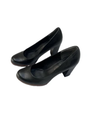 Svarta pumps från Marco Tozzi - Klassiska svarta pumps från Marco Tozzi med rund tå och bred klack. Skorna är tillverkade i svart läder och har en stilren design som passar till många olika looks. Storlek 37. Endast testade inomhus.