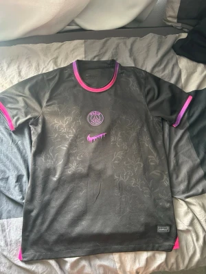 Svart PSG x Nike t-shirt med rosa detaljer - Snygg svart PSG t-shirt från Nike med diskret blommönster i tyget. Rosa detaljer vid krage och ärmslut, samt rosa PSG-logga och Nike swoosh med drip-effekt på bröstet. Perfekt för dig som gillar streetwear och fotbollsinspirerad stil.