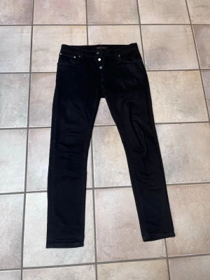Svarta jeans från Nudie Jeans - Snygga svarta raka jeans från Nudie Jeans med klassisk femficksmodell och silverfärgade knappar. Jeansen har en smal passform och är tillverkade i ett stretchigt material för extra komfort. Perfekta till en avslappnad streetwear-look.