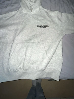 Grå Essentials Fear of God hoodie - Säljer en ljusgrå hoodie från Essentials Fear of God med svart logga på bröstet. Hoodien har stor huva och en klassisk magficka. Tillverkad i mjukt och bekvämt material, perfekt för en avslappnad streetwear-look.