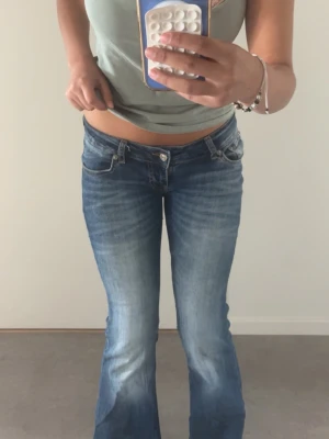 Blå bootcut jeans med låg midja - Säljer ett par blå bootcut jeans med låg midja och klassisk femficksmodell. Jeansen har en snygg tvätt med slitningar framtill och är tillverkade i denim med lätt stretch. Perfekta för en trendig och avslappnad look.