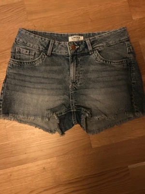 Blå jeansshorts från Lindex - Snygga blå jeansshorts från Lindex med rå kant nertill och klassisk femficksdesign. Shortsen har dekorativa sömmar på bakfickorna och en knappgylf framtill. Perfekta för sommaren och ger en avslappnad vibe.