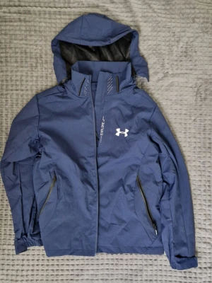 Blå regnjacka med huva Under Armour - Blå regnjacka från Under Armour med huva och dragkedja framtill. Jackan har två snedställda fickor med dragkedja och vit logotyp på bröstet. Insidan är fodrad med mesh för extra komfort och ventilation. Tillverkad i vattenavvisande material.
