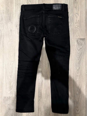 Nudie Jeans Lean Dean Svarta Selvage W29/L28 - Slim Tapered Fit - Nudie Jeans Lean Dean ”Dry Black Selvage” svarta selvage-jeans med slim tapered-passform, normalhög midja och avsmalnande ben, storlek W29/L28, midja 40 cm, ytterbenslängd 92,5 cm, benöppning 15,5 cm, modellen är 185cm & väger 65kg, i fint skick.