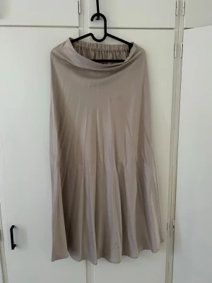 Beige långkjol i satin - Säljer en beige långkjol med glansig satinlook och elastisk midja. Kjolen har en mjuk och följsam passform som ger ett snyggt fall. Perfekt för dig som gillar stilrena och enkla plagg.  Har en liten fläck men går förmodligen bort med tvätt.