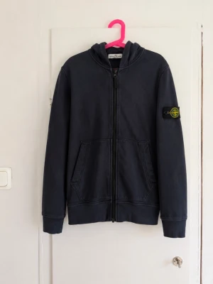 Mörkblå Stone Island Junior zip hoodie med den klassiska Stone Island-badgen på ärmen 🧭 - Snygg och tidlös modell med hel dragkedja, huva och två fickor framtill. Klassisk mörkblå/navy färg som passar till det mesta. Hoodien är använd och har en snyggt tvättad/faded look. Bra begagnat skick, se bilder för skick och detaljer. • Stone Island Junior • Mörkblå/navy • Avtagbar Stone Island-badge på ärmen • Full zip • Huva • Två framfickor Skriv gärna för mått eller fler bilder!