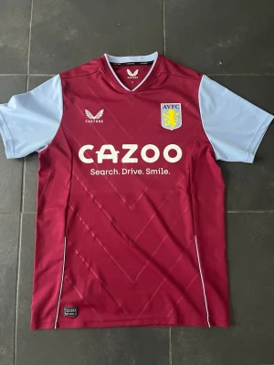 Aston Villa matchtröja Archer 35 - Snygg Aston Villa matchtröja från Castore med Archer och nummer 35 på ryggen. Tröjan är vinröd med ljusblå ärmar, klubbmärke och sponsortryck på bröstet. Tillverkad i lätt och ventilerande material, perfekt för fotboll eller träning.