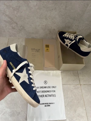 Blåa Golden Goose sneakers med stjärna - Säljer ett par snygga sneakers från Golden Goose i blå mocka med vit stjärndetalj på sidan och vita skosnören. Skorna har vit sula och hälparti samt klassisk låg profil. Perfekta för dig som gillar streetstyle och unika detaljer.