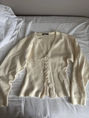 Krämvit kofta med knappar - Snygg krämvit kofta med v-ringning och dekorativa knappar framtill. Koftan har lång ärm och är tillverkad i ett mjukt, stickat material som ger en clean och stilren look. Perfekt att matcha med jeans eller kjol.