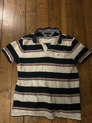 Randig pikétröja från Tommy Hilfiger - Randig pikétröja från Tommy Hilfiger i marinblått, vitt och röda detaljer. Klassisk krage, knappar vid halsen och korta ärmar. Tillverkad i mjuk bomull för en bekväm känsla. Perfekt för en avslappnad och stilren look.