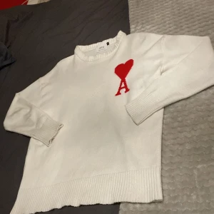 Vit stickad tröja från Ami med rött hjärta - Vit stickad tröja från Ami med ett stort rött hjärta och bokstaven A på bröstet. Tröjan har ribbade muddar vid ärmslut och nederkant, rund hals och långärmad passform. Perfekt för dig som gillar clean stil med en twist.