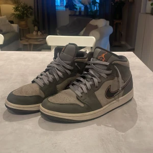 Nike Air Jordan 1 Mid grå/svart  - Nike Air Jordan 1 Mid i grått och svart mocka och skinn, med orange detaljer på tungan och swoosh. Klassisk Air Jordan-logga på sidan, svart yttersula och grå snörning. Snygg och tidlös modell som passar till streetwear och sneakersamlingen. 😇 Boxen till skorna får man tyvärr inte med.  Ny pris 1900, vårt pris 1100😅