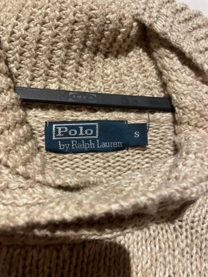 Beige polotröja Polo Ralph Lauren S - Stickad beige polotröja från Polo Ralph Lauren i storlek S. Tröjan har en bred, ribbad polokrage med knappdetalj och ett stort amerikanskt flaggmotiv i blått, vitt och rött framtill. Mjukt material och ribbade muddar vid ärmslut och nederkant.