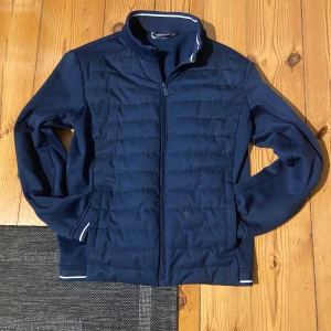 Marinblå quiltad jacka från Marine - Snygg marinblå jacka från Marine med quiltad front och hel dragkedja. Jackan har ribbade muddar med vit rand, ståkrage och diskret logga på bröstet. Perfekt för dig som gillar stilren och sportig look.