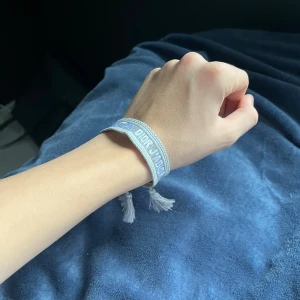 Grått Christian Dior Armband - Snyggt grått friendship-armband från Christian Dior med broderad logga och två dekorativa tofsar. Armbandet är justerbart med knytning och har en stilren, modern look som passar perfekt till din outfit.