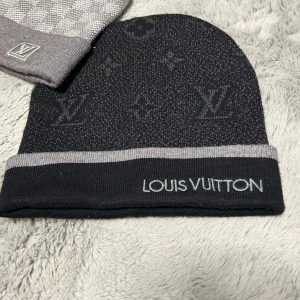 Svart mössa från Louis Vuitton - Snygg svart mössa från Louis Vuitton med grå kant och broderad logga framtill. Mössan har diskret LV-monogram och klassiska Louis Vuitton-blommor i mönstret. Tillverkad i mjukt stickat material som håller dig varm och stilren.