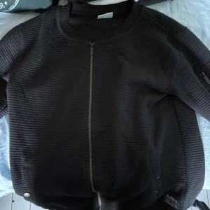 Svart bomberjacka från COPE - Säljer en svart bomberjacka från COPE med ribbad struktur och dragkedja framtill. Jackan har fickor med tryckknappar och en extra ficka på ärmen. Perfekt för dig som gillar en clean och stilren look.