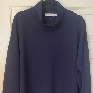 Marinblå klänning / lång tunika  Carin Wester  - Snygg marinblå klänning / lång tunika från Carin Wester i modellen Julie. Klänningen har lång ärm, hög krage och en rak, loose passform. Längd i ärm: 1 meter. Materialet är mjukt och stretchigt med 81% polyester, 14% viskos och 5% elastan. 