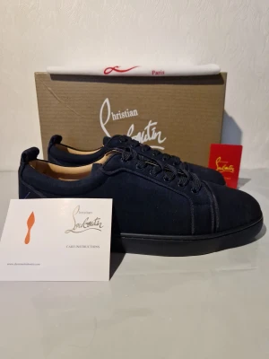 Mörkblå sneakers från Christian Louboutin 43 - Snygga mörkblå sneakers från Christian Louboutin med klassisk röd sula och broderad logga på hälen. Skorna har rund tå, snörning och är tillverkade i mocka med lyxig känsla. Perfekt för dig som vill sticka ut med exklusiva detaljer och stilren design.