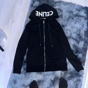 Svart hoodiejacka från Celine - Svart hoodiejacka från Celine med stor vit logga på huvan. Jackan har dragkedja framtill, två fickor och justerbara snören vid huvan. Tillverkad i mjuk bomull för skön känsla och stilren look. Perfekt för dig som gillar exklusiva streetwear-vibbar.