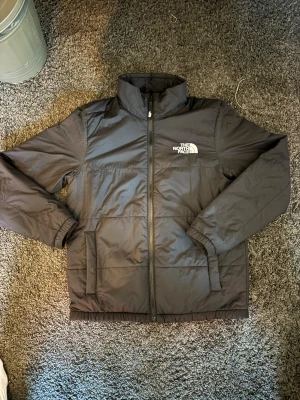 Svart pufferjacka från The North Face - Svart pufferjacka från The North Face i storlek M. Två sidofickor och dragkedja framtill. Loggan är tryckt på både bröstet och ryggen. Materialet är syntet och jackan är perfekt för kyliga dagar.