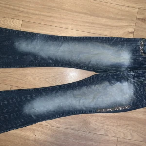 Blå bootcut jeans  - Säljer ett par blå bootcut jeans med coolt blekt mönster och metalldetaljer som ger en edgy vibe. Perfekt för dig som gillar statement-jeans med unik look. Midjemåttet rakt över =37/38 cm, Innerbenslängden= 82 cm, skulle säga jeansen passar xs/s