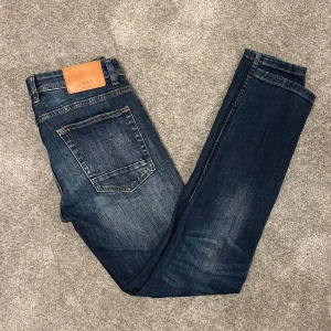 Mörkblå jeans från Smog - Snygga mörkblå jeans från Smog med skinny passform och klassisk femficksdesign. Jeansen har slitningar och tvättade detaljer på låren och knäna, samt en brun läderpatch med logga bak i midjan. Perfekt för dig som gillar en smal siluett.