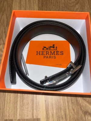 Svart skärp med H-spänne - Exklusivt svart bälte från med klassiskt H-spänne i silverfärgad metall. Bältet är tillverkat i slätt skinn och levereras i den ikoniska orange boxen. 