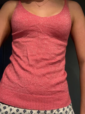 Glittrig rosa linnetopp med smala axelband - Säljer en snygg rosa linnetopp med glittrigt material och smala axelband. Toppen har en v-ringad hals och ribbad kant nedtill. Perfekt för dig som vill sticka ut med lite extra shimmer. Passformen är normal och toppen är ärmlös.