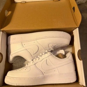 Nike Air Force 1 helvita sneakers - Nike Air Force 1 sneakers i helvitt skinn med klassisk låg siluett och rund tå. Snygga detaljer som perforering på tån, snörning och tydlig Nike-logga på hälen. Perfekt för dig som gillar clean och stilren look. Ny pris 1300 