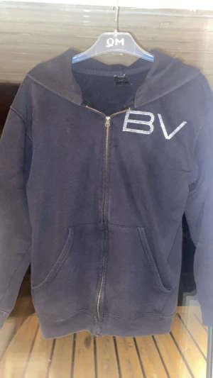 Svart hoodie med strass från Bvaldi - Svart hoodie från Bvaldi med strassdetaljer på bröstet och ryggen. Framtill finns BV i strass och på ryggen syns änglar och BVALDI i strass. Tröjan har huva, dragkedja och två fickor framtill. Perfekt för dig som gillar bling och streetstyle. Passar på mig som är storlek S. 